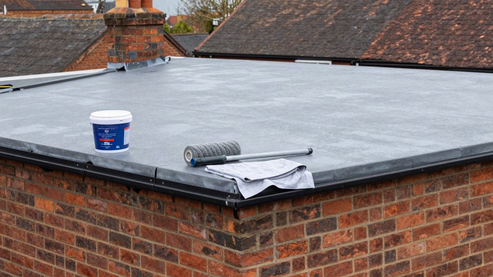 A grey felt flat roof on a UK brick garage part-coated with elastomeric roof sealant, with a tub, a roller on a pole and a roll of flashing tape beside it