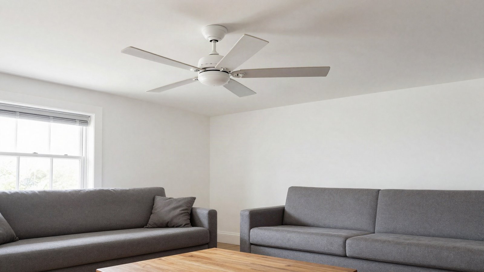 A white five-blade ceiling fan mounted on a painted ceiling above a bright British living room with a sofa and open sash window