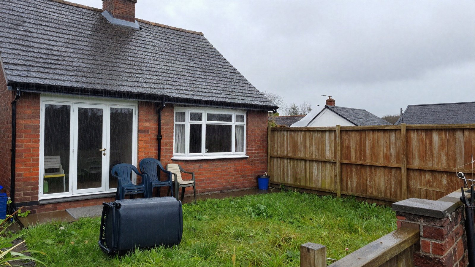 Wind and heavy rain battering the rear of a British semi-detached house, with garden furniture stacked against the wall and a bowing timber fence panel