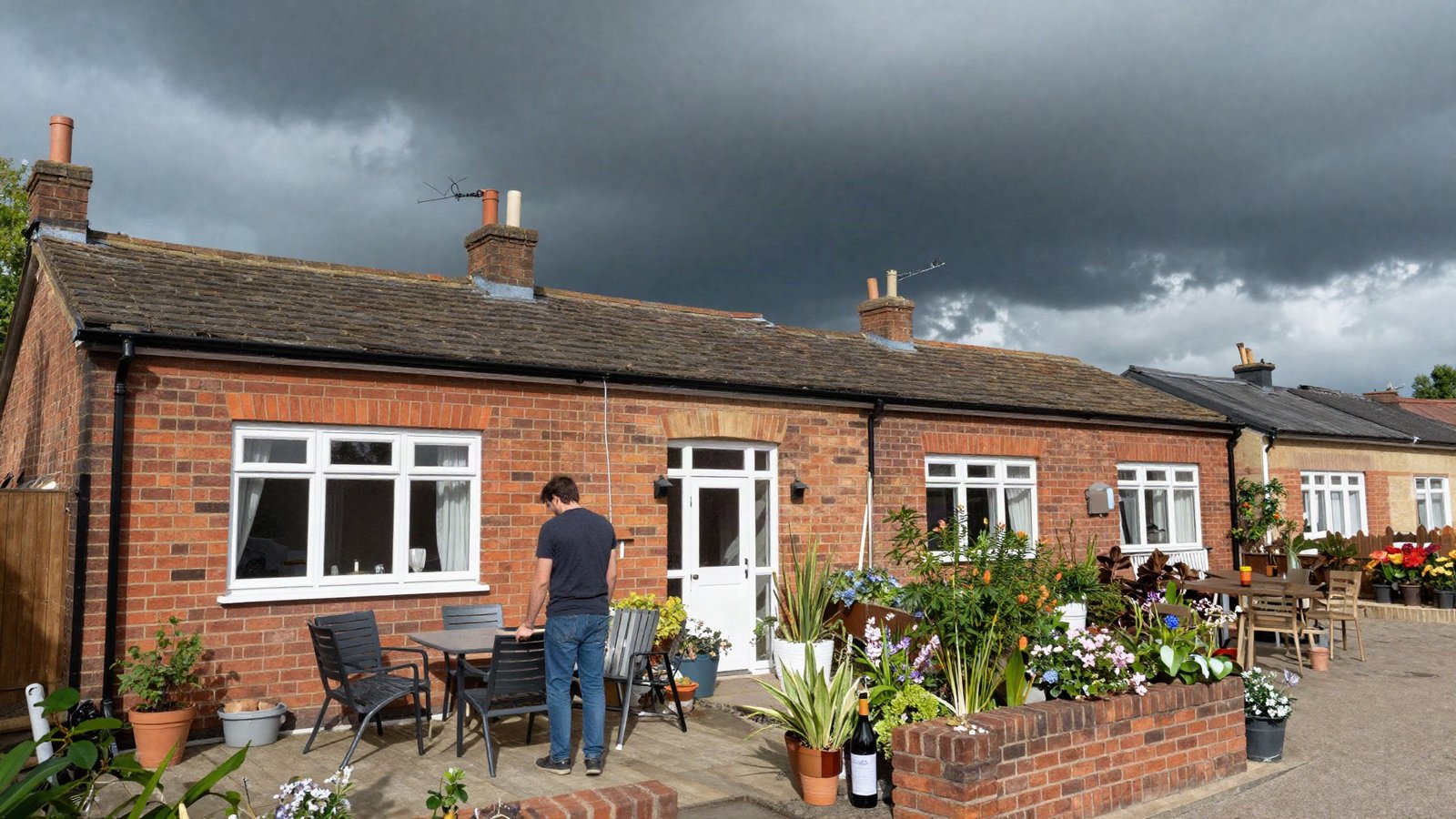 Homeowner securing garden furniture outside a brick UK terraced house as dark storm clouds gather overhead