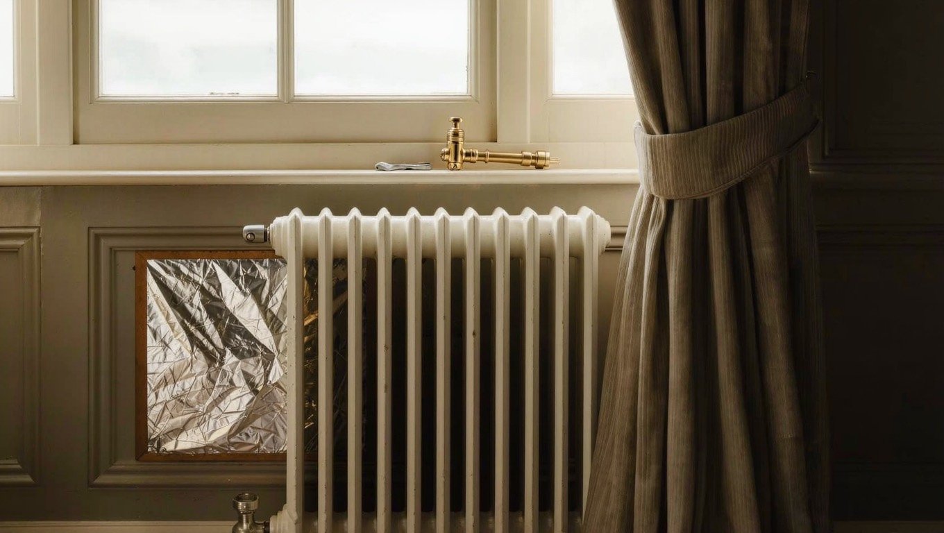 A white column radiator beneath a sash window in a UK living room, with the curtain drawn back and soft autumn daylight